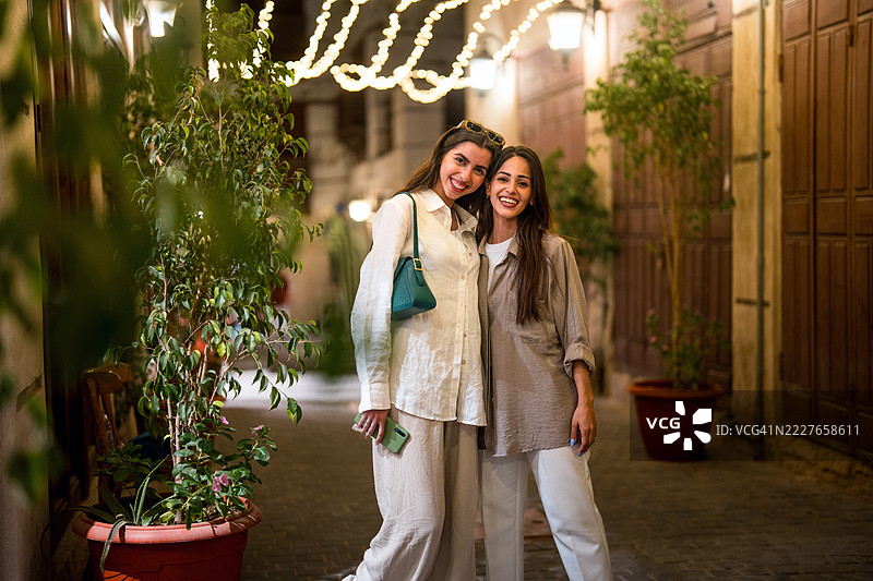 两位微笑的女性在夜晚的阿尔巴拉德灯光巷道中与盆栽植物合影图片素材
