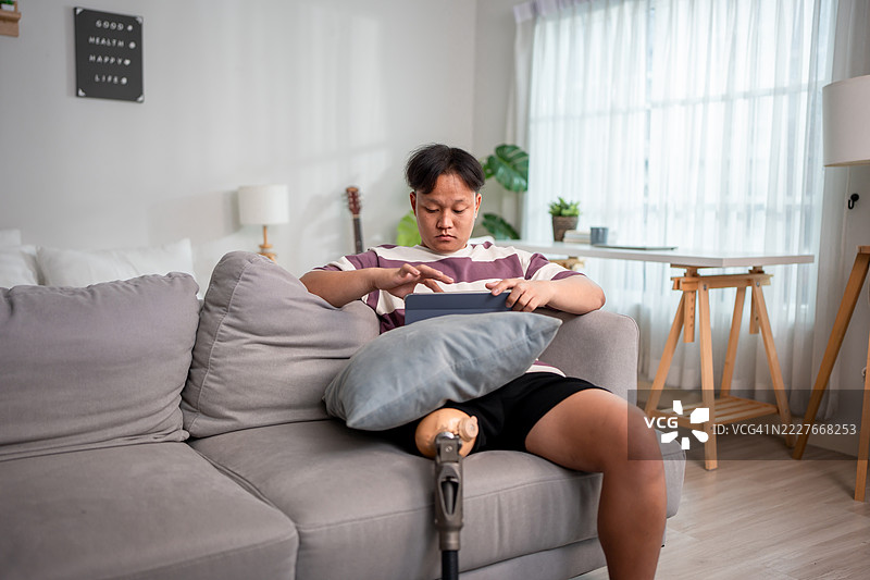 亚洲男子在家中的沙发上使用数字平板电脑，身穿义肢。吸引人的年轻男性截肢者穿着腿部支架，享受在舒适的家中度过闲暇时光，放松身心，进行康复。图片素材