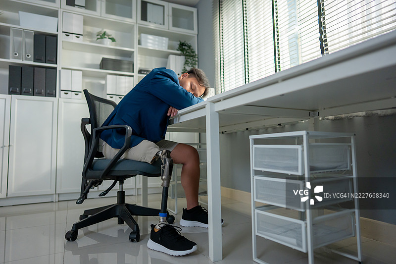 亚洲年轻男子在办公室的工作桌上睡觉，腿部有假肢。吸引人的男性截肢者穿着腿部支架，感到疲惫和困倦，在专业的工作环境中休息。图片素材