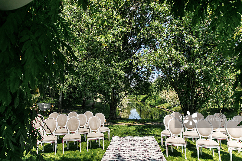 优雅的户外婚礼布置，花卉拱门和白色椅子位于阳光明媚的郁郁葱葱的花园中。图片素材