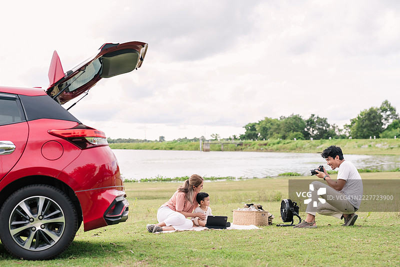 父亲在湖边用平板电脑为家人拍照，旁边停着一辆红色SUV。图片素材