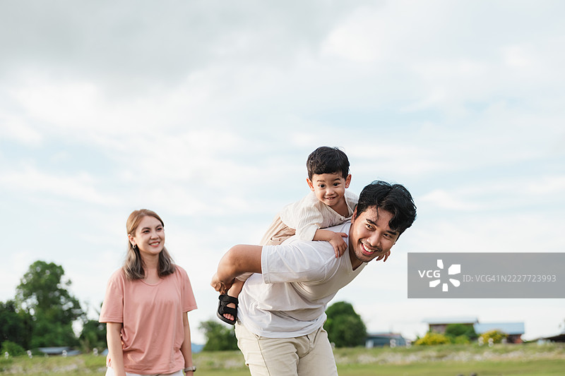 亚洲家庭在大自然中享受户外乐趣。父亲给年轻的儿子背着，母亲在旁微笑，捕捉到幸福、爱与快乐的亲密时光，在开阔的草地上。图片素材