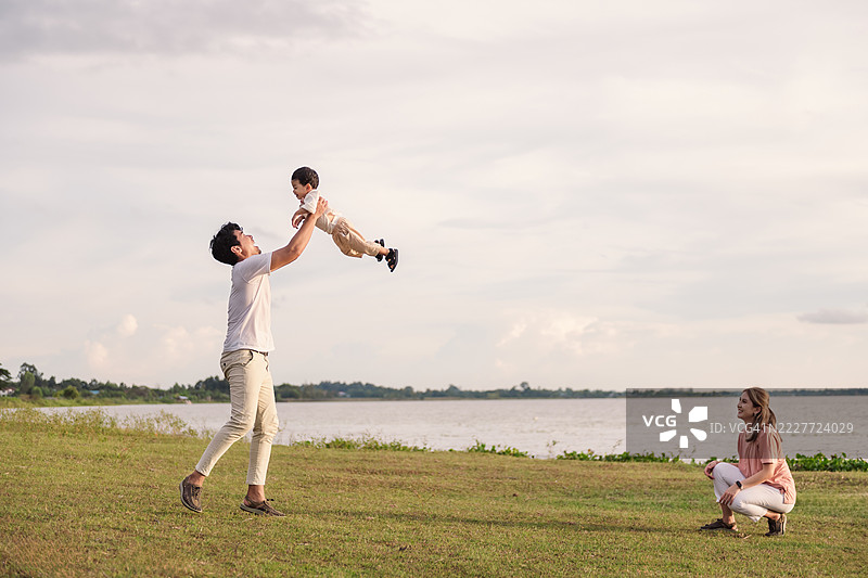 父亲在河边与儿子玩耍，把孩子高高举起，母亲在旁微笑。快乐的亚洲家庭，亲情、信任和在大自然中愉快的亲密关系。图片素材