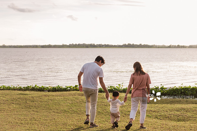 亚洲家庭在日落时分享受河边的户外活动。父母手牵着儿子，走在大自然中，微笑着在宁静快乐的时刻中增进感情。图片素材