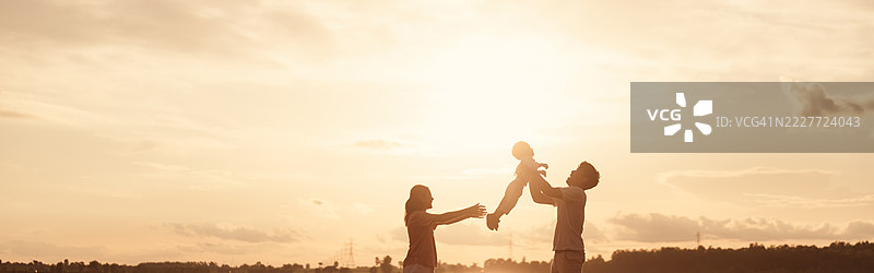 亚洲家庭在日落时享受户外时光。父亲将孩子高高举起，母亲伸出手，营造出一个温馨的爱、关怀和团聚的时刻，沐浴在金色的光芒中。图片素材