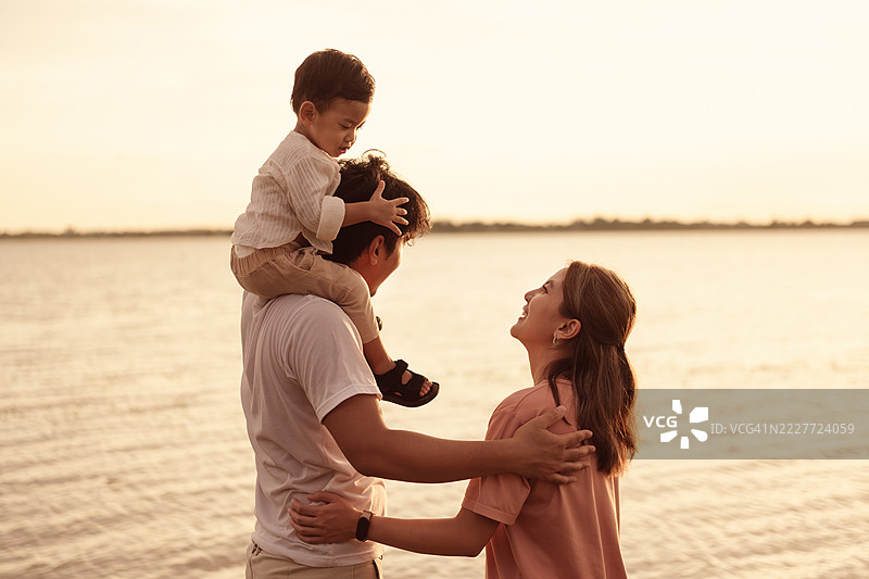 快乐的亚洲家庭在日落时分河边亲密相处。父亲肩上扛着孩子，母亲温暖地微笑，捕捉到爱、快乐和团聚的放松时刻。图片素材