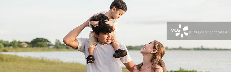 快乐的亚洲家庭在河边享受户外的美好时光。父亲给儿子骑肩膀，母亲微笑着，捕捉到大自然中快乐而温馨的瞬间。图片素材
