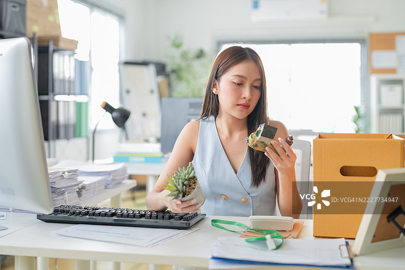 一位亚洲女商人在现代办公室的背景下，悲伤地将自己的物品装进箱子里，展现了她被解雇后的艰难时刻。图片素材