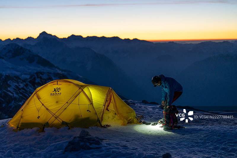 中年登山者在雪-covered阿尔卑斯山景区的黄帐篷附近准备装备，日出时分。图片素材