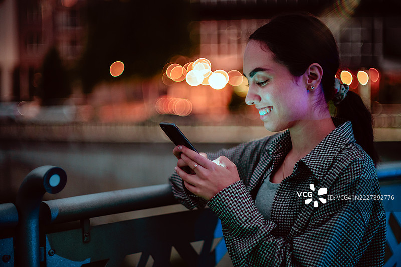 一位微笑的年轻女性在城市的晚上用手机发信息图片素材