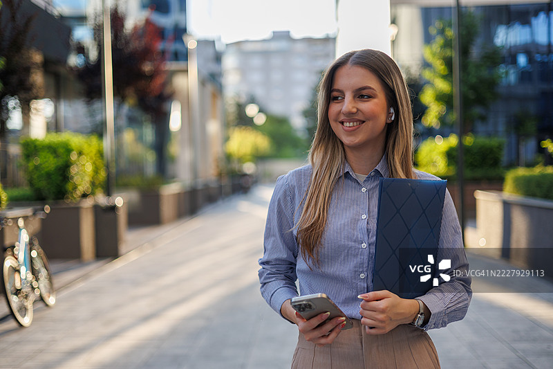 微笑的女商人在城市街道上走着，一边使用智能手机，一边通过无线耳机享受音乐。图片素材
