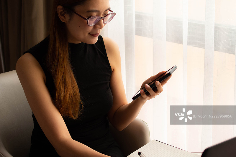 一位中年千禧一代的东南亚女性在家中无缝使用智能手机和笔记本电脑——通过用户友好的应用程序和平台检查余额、进行交易和管理预算。图片素材