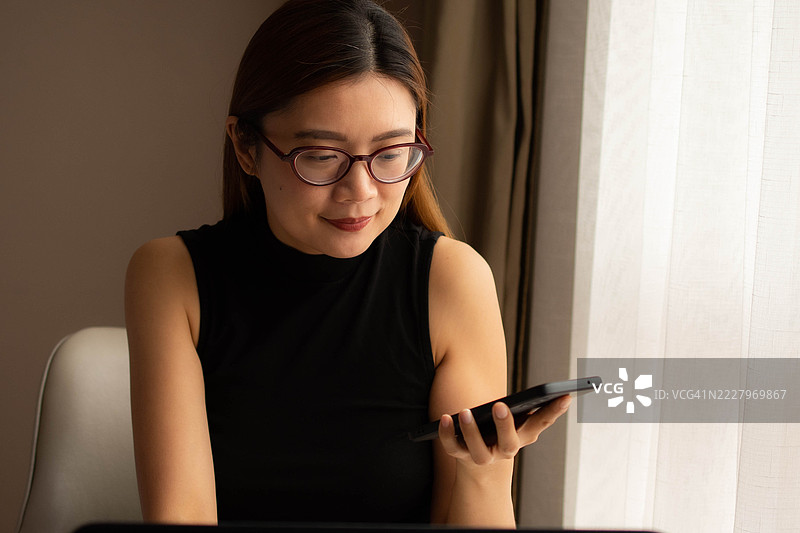 一位中年千禧一代的东南亚女性在家中无缝使用智能手机和笔记本电脑——通过用户友好的应用程序和平台检查余额、进行交易和管理预算。图片素材