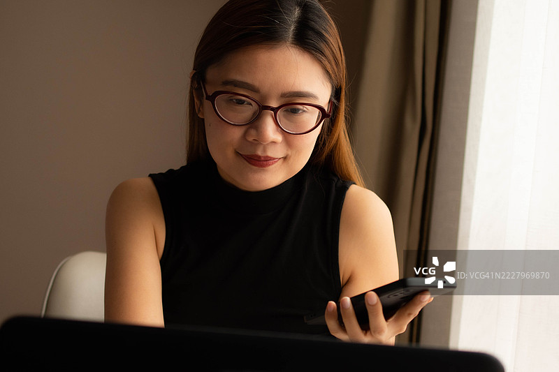 一位中年千禧一代的东南亚女性在家中无缝使用智能手机和笔记本电脑——通过用户友好的应用程序和平台检查余额、进行交易和管理预算。图片素材