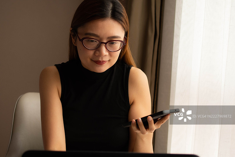 一位中年千禧一代的东南亚女性在家中无缝使用智能手机和笔记本电脑——通过用户友好的应用程序和平台检查余额、进行交易和管理预算。图片素材