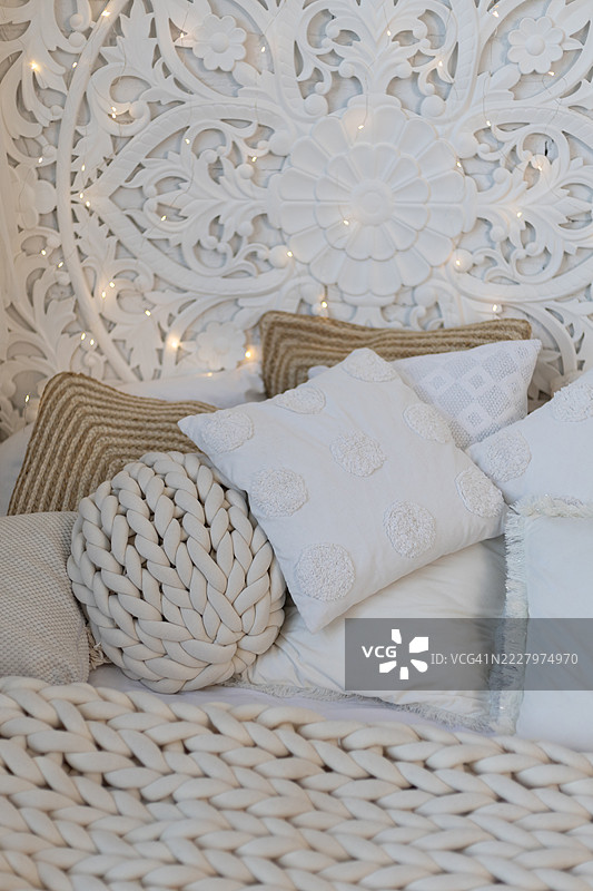 雕刻的床头板上挂着 fairy lights，柔软的床铺上铺着厚实的针织毛毯，旁边有一个编织的圆形靠垫和几个枕头。图片素材