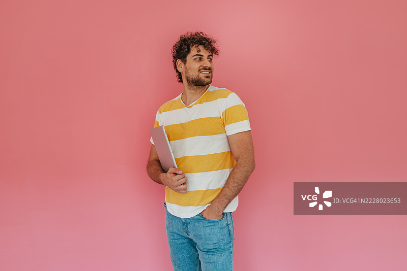穿着休闲服的快乐年轻男子，卷发，站在鲜艳的粉色墙壁前，展现出快乐和自信。图片素材