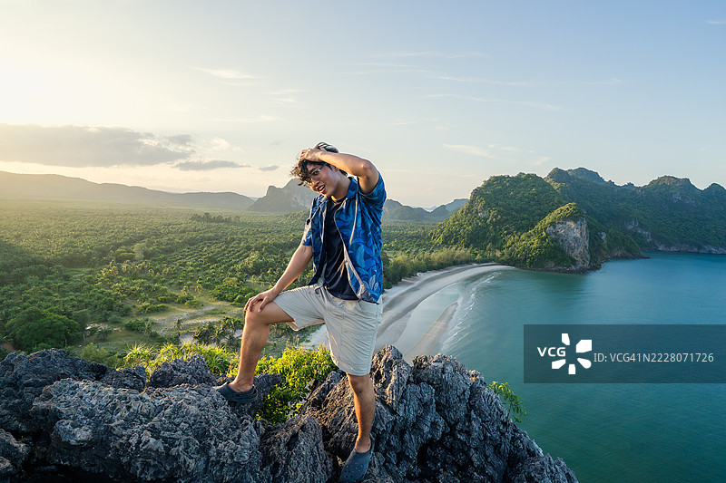 穿着休闲服的旅行者自信地站在岩石悬崖上，欣赏着热带海岸线和郁郁葱葱的风景，沐浴在金色时光的光辉中。图片素材