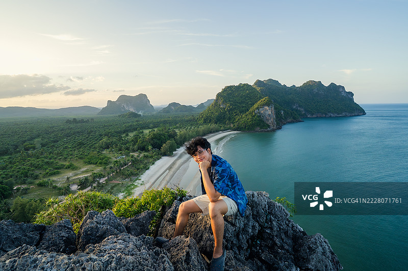 穿着休闲服的旅行者自信地站在岩石悬崖上，欣赏着热带海岸线和郁郁葱葱的风景，沐浴在金色时光的光辉中。图片素材