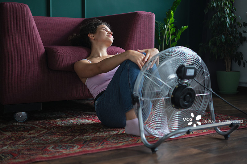 微笑着满足的女人坐在电风扇旁，享受着炎热夏日的新鲜空气，度过热浪，生活在没有空调的公寓里。图片素材