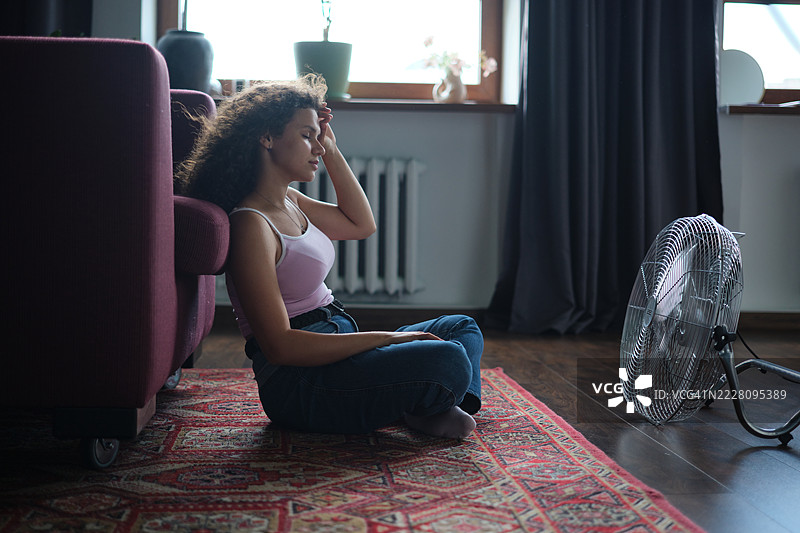 年轻疲惫的卷发女性坐在地板上，面前有电风扇，享受着清凉的空气，忍受着夏季的炎热，住在没有空调的公寓里。图片素材