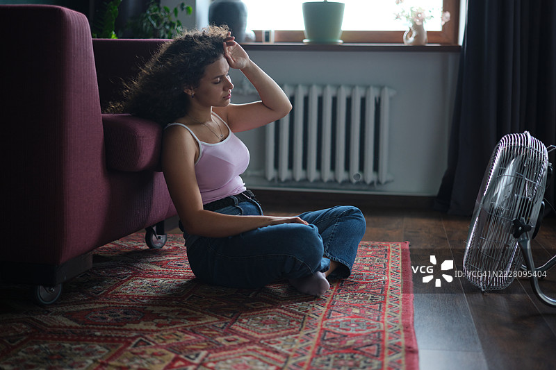 疲惫不堪的年轻女性在夏季炎热中感到不适，坐在电风扇前，居住在没有空调的公寓里。图片素材