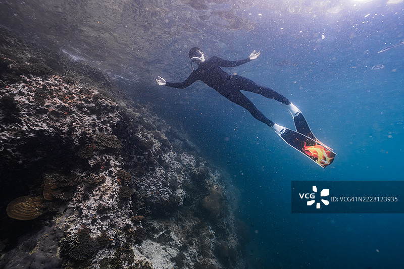 一名穿着黑色潜水服和火焰图案鳍的男性自由潜水员在清澈的热带海洋水域上方游泳，周围是珊瑚礁。图片素材