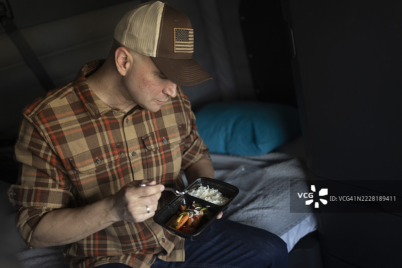 一位饥饿的卡车司机在卡车里吃着他从微波炉加热的健康米饭和蔬菜餐。图片素材