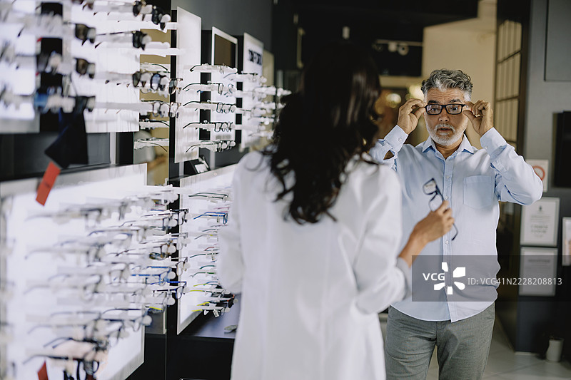 年轻女性验光师在现代眼镜店帮助一位老年男性客户选择眼镜框。图片素材