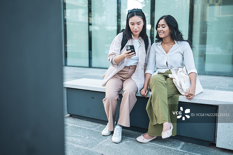 两个年轻女性正在使用智能手机，坐在长椅上。图片素材