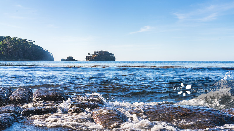 波浪拍打在岩石海岸上，远处的悬崖在晴朗的天空下。图片素材