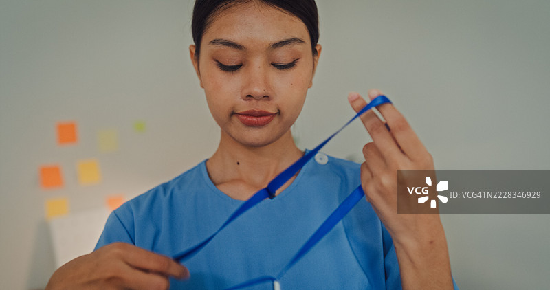 年轻亚洲女性护士在卧室里穿着米色毛衣，身穿蓝色护士服的特写。护士生活方式概念。图片素材