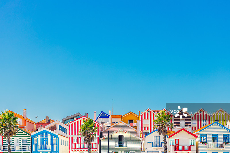 色彩斑斓的海滩小屋位于科斯塔诺瓦，展现出鲜艳的红色、蓝色、黄色和绿色图案，周围有棕榈树和明亮的蓝天。图片素材