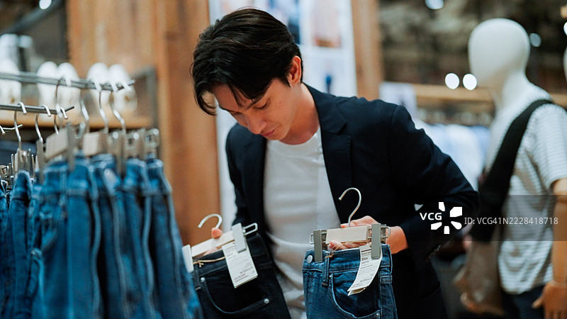 年轻的亚洲男子在购物中心比较黑色和蓝色牛仔裤，准备购买一条。图片素材