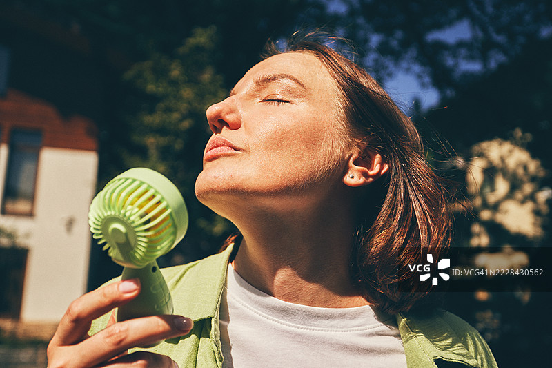 女人在阳光明媚的天气中，享受着户外新鲜空气，手持便携风扇。图片素材