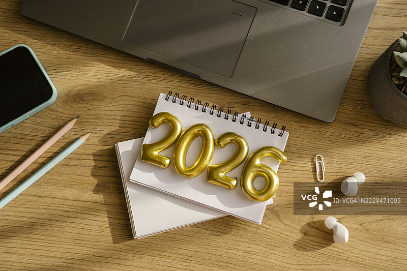 木质工作桌上的笔记本，金色数字2026。为2026年制定新年计划。笔记本、笔记本电脑、手机、铅笔。图片素材