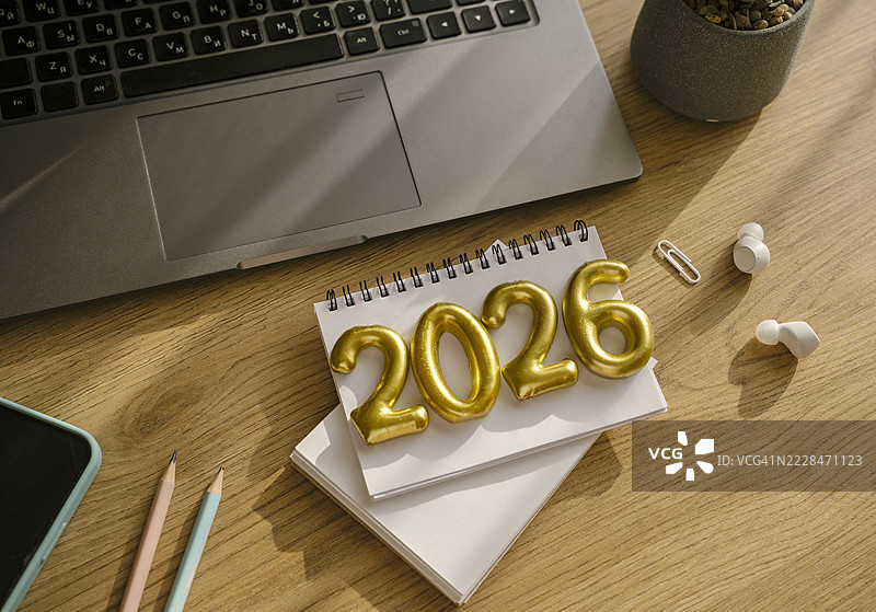 木质工作桌上的笔记本，上面写着金色数字2026。为2026年制定新年计划。笔记本、笔记本电脑、手机、铅笔。图片素材