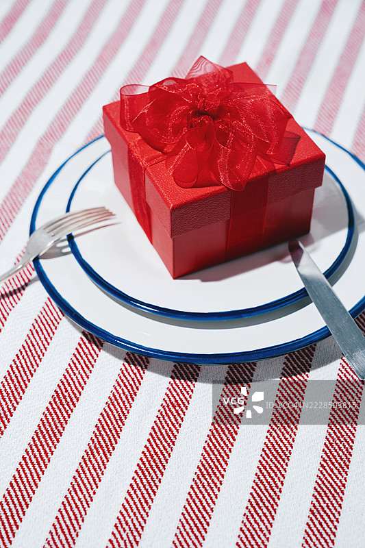 红色礼品盒，上面系着丝带，放在带餐具的盘子上。桌子，条纹桌布。俯视图。样机。图片素材