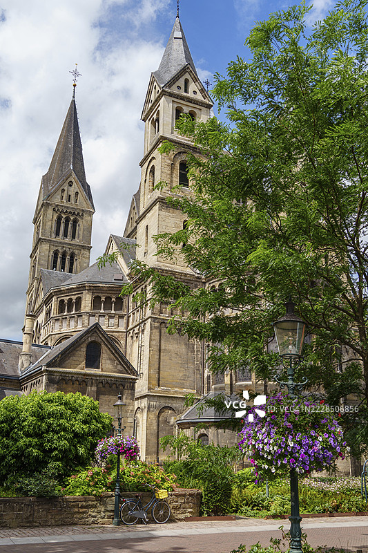 历史悠久的教堂,哥特式塔楼被郁郁葱葱的树木和盛开的花朵环绕,天空多云,位于荷兰林堡省的罗ermond。图片素材