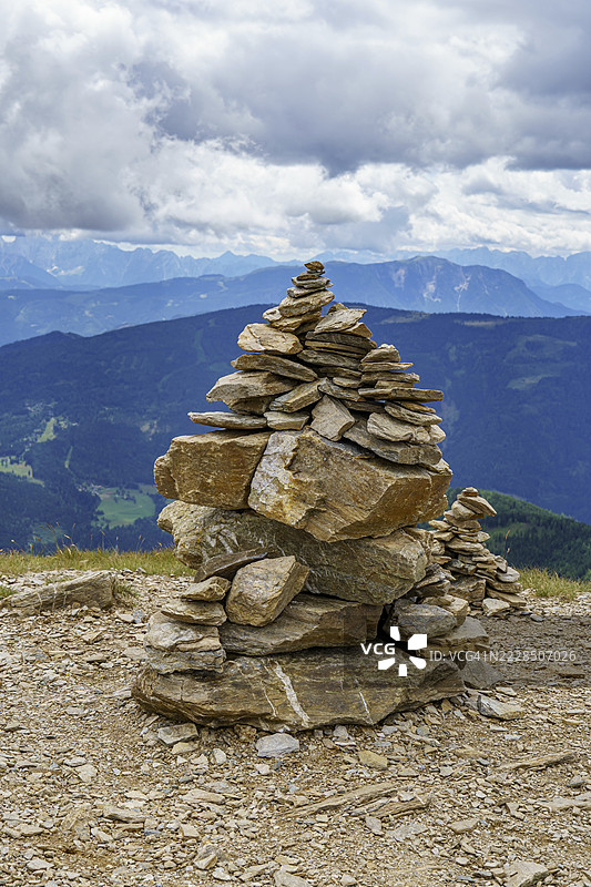 岩石地面上的石堆，背景是云朵和山脉风景，地点：奥地利卡林西亚州的巴德克莱因基希姆图片素材