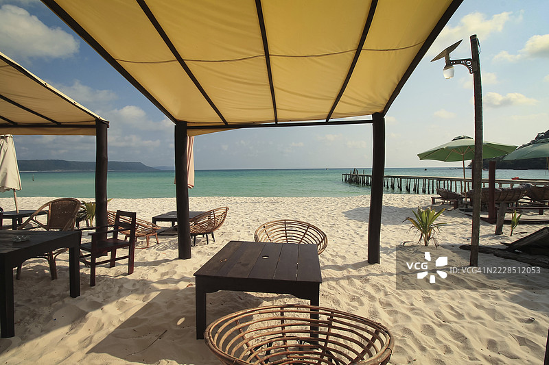 在柬埔寨西哈努克市的考荣山洛姆公共海滩上，沙滩上有遮阳篷下的桌子，背景是平静的海洋。图片素材