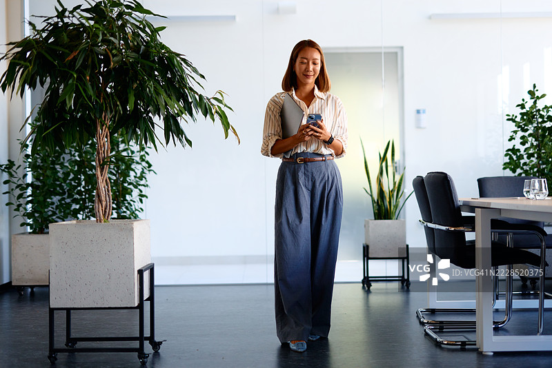 年轻的亚洲女商人在现代办公室中使用智能手机，周围有植物。图片素材