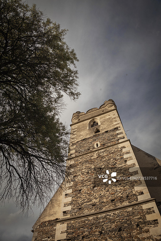 从近距离视角看，一棵树旁边的一座教堂塔楼，背景是多云的天空。瓦豪瓦尔德维尔特圣迈克尔教堂，下奥地利，奥地利。图片素材