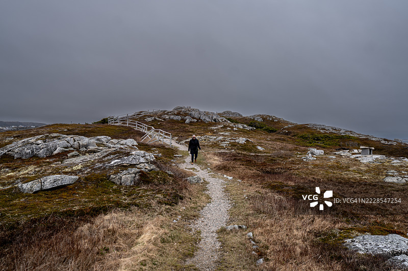 一名女性背对着镜头走在步道上，朝着暴风雨中的玫瑰白灯塔前进，地点为加拿大纽芬兰和拉布拉多省的玫瑰白港。图片素材