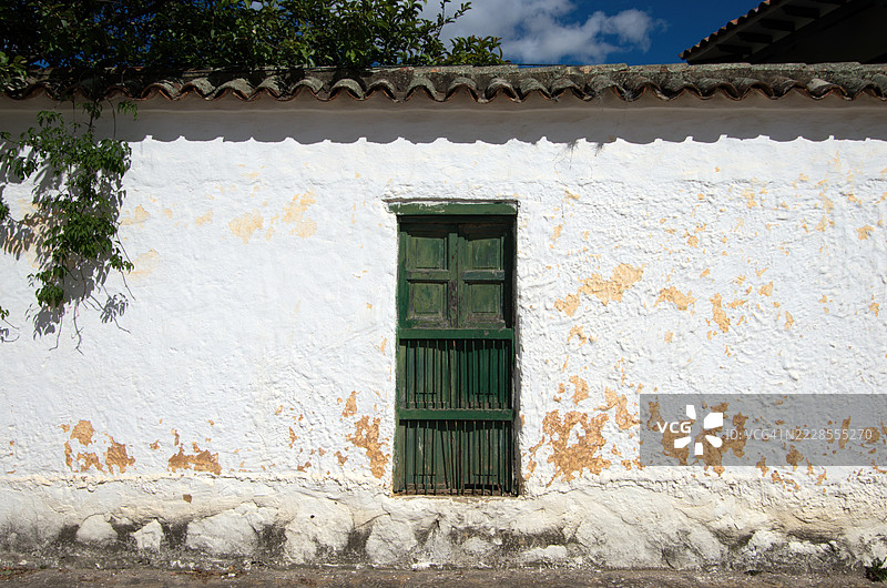质朴的白washed和风化的殖民时代建筑，带有纹理的外立面，绿色木制百叶窗，配有垂直安全栅栏和陶土瓦屋顶。图片素材