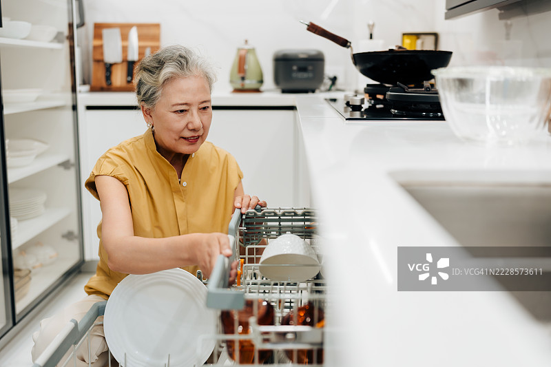 亚洲老年女性蹲下在厨房使用洗碗机。图片素材