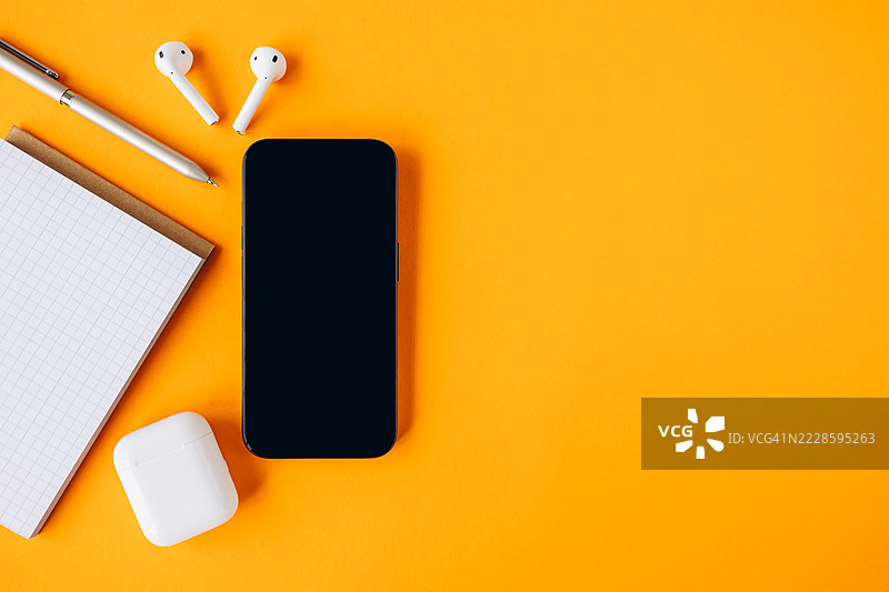 平铺的智能手机，白色空屏，无线耳机，钢笔和记事本，背景为鲜艳的黄色。现代工作空间概念图片素材