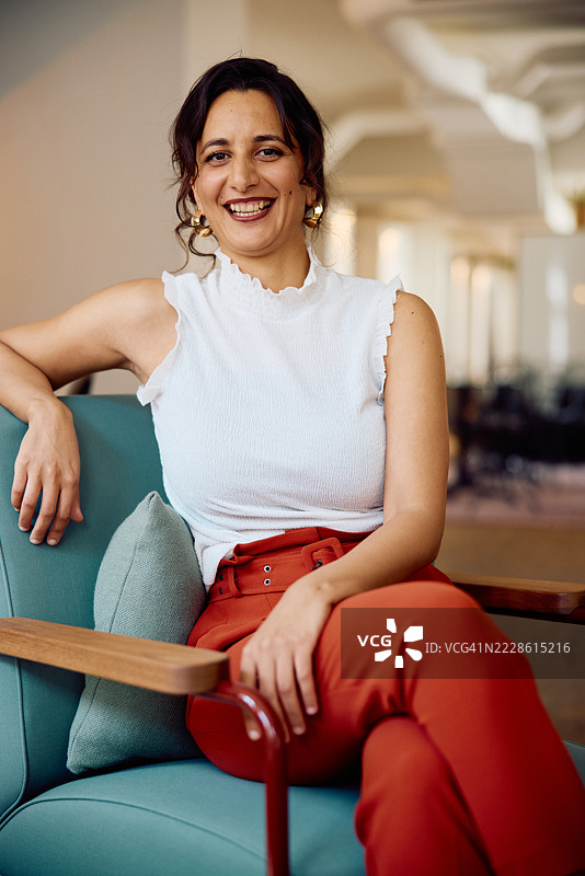 一位快乐的职业女性坐在椅子上，双腿交叉，背景是现代办公室的室内环境。图片素材