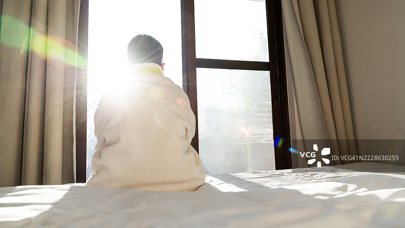 一个青少年男孩背对着坐在卧室的床上，独自望向窗外。图片素材