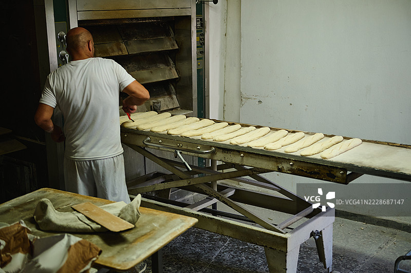 在传统面包店中，面包师正在准备面包坯子，旁边有烤箱。图片素材